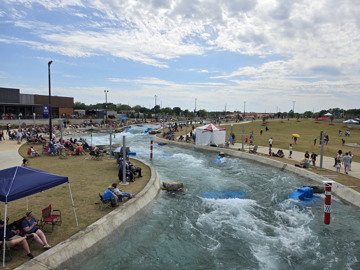 A wide view of an outdoor artificial whitewater course designed by a skilled whitewater engineer, with people sitting and standing along the grassy sides, tents for shade, and partly cloudy skies overhead. Blue rafts float on the churning water.