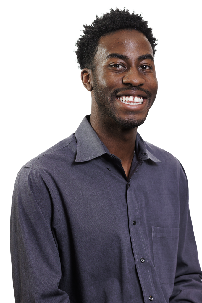 A young man with short curly hair and a beard is smiling broadly. He is wearing a button-up, long-sleeve gray shirt and is posed against a plain white background, showcasing the approachable side of a Colorado engineering professional.