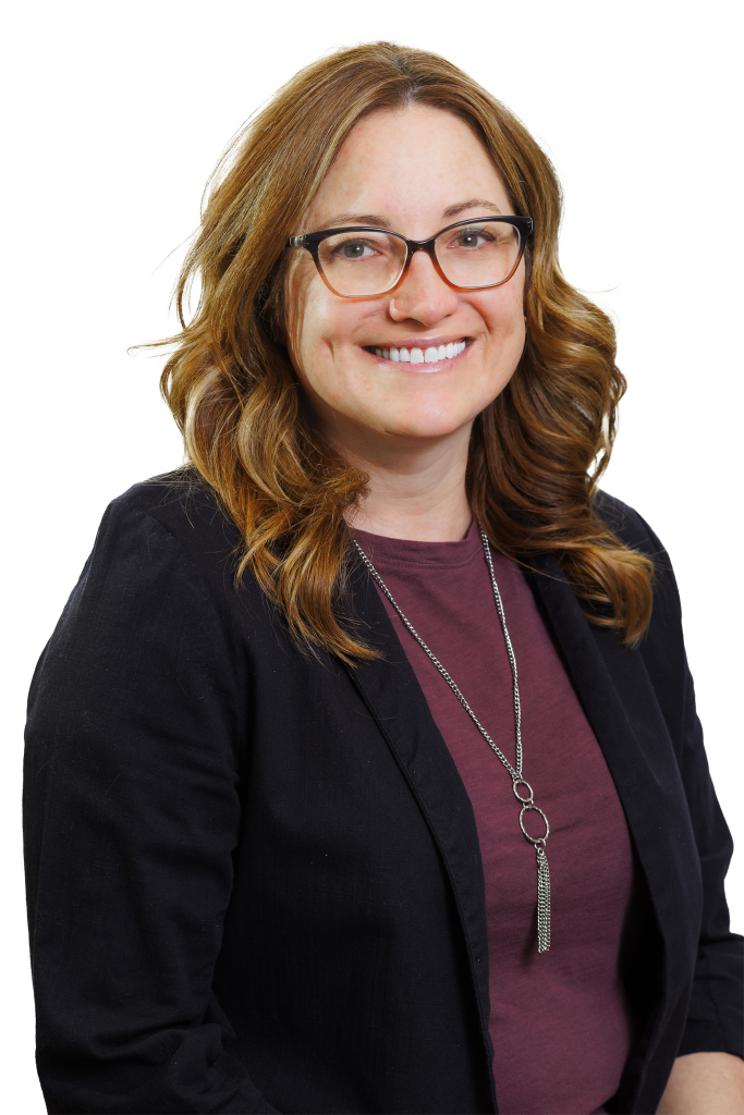 A woman with wavy brown hair and glasses smiles at the camera. She is a structural engineer in a black blazer, maroon top, and long silver necklace. The background is white.