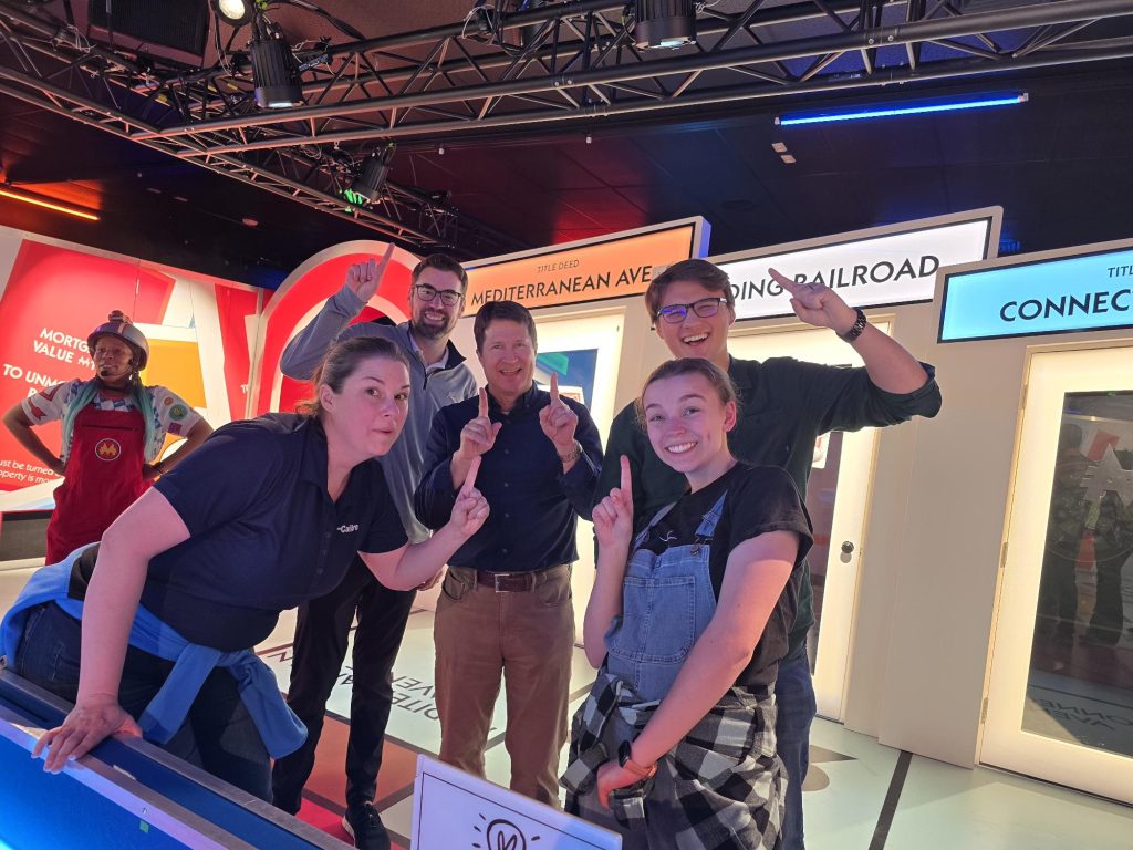 Five people stand indoors, smiling and holding up one finger each in front of colorful Monopoly-themed signs. Among them is a Denver engineer, adding excitement to what looks like a game or competition victory.