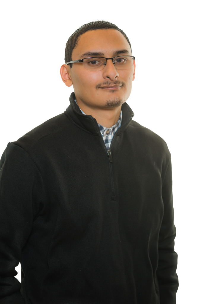 A young man with short hair, glasses, and a mustache is wearing a black zip-up sweater over a blue plaid shirt. He stands with a neutral expression, embodying the professional look of a civil engineering or structural engineer.