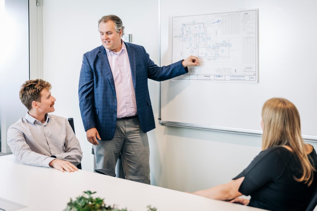 A Denver engineer in a suit stands at a whiteboard, pointing to a technical drawing while two colleagues, one male and one female, sit at a table and listen attentively.
