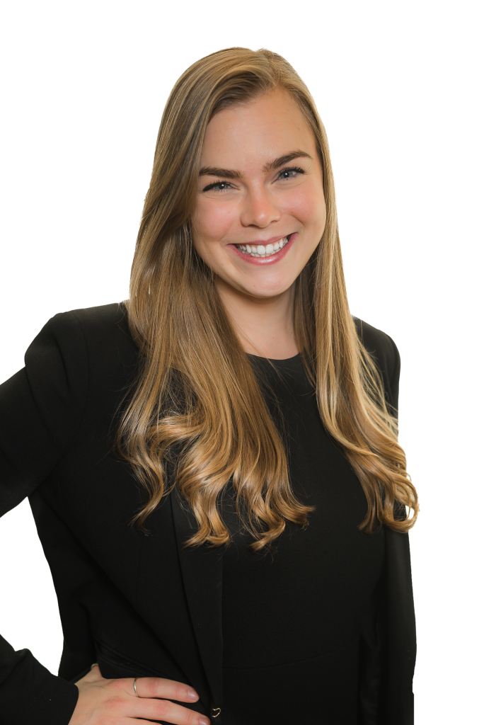 A smiling woman with long, wavy blonde hair wearing a black blazer and black top stands with one hand on her hip, confidently embodying the professionalism of a structural engineer against a plain background.