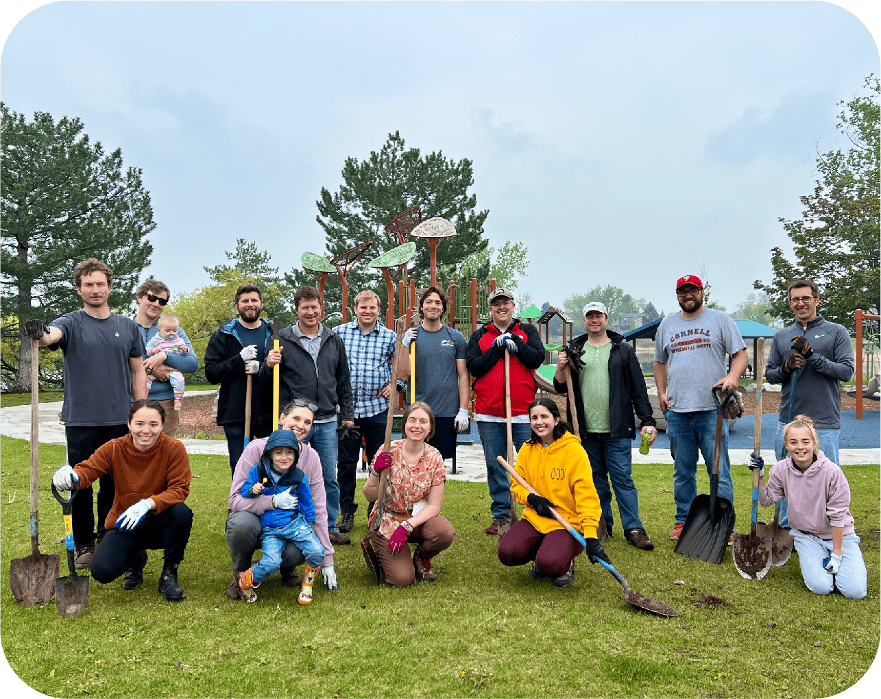 A group of people, including a Denver engineer, pose outdoors on grass with shovels and gardening tools. Some kneel or crouch in front while others stand behind. Trees and playground equipment are visible in the background.