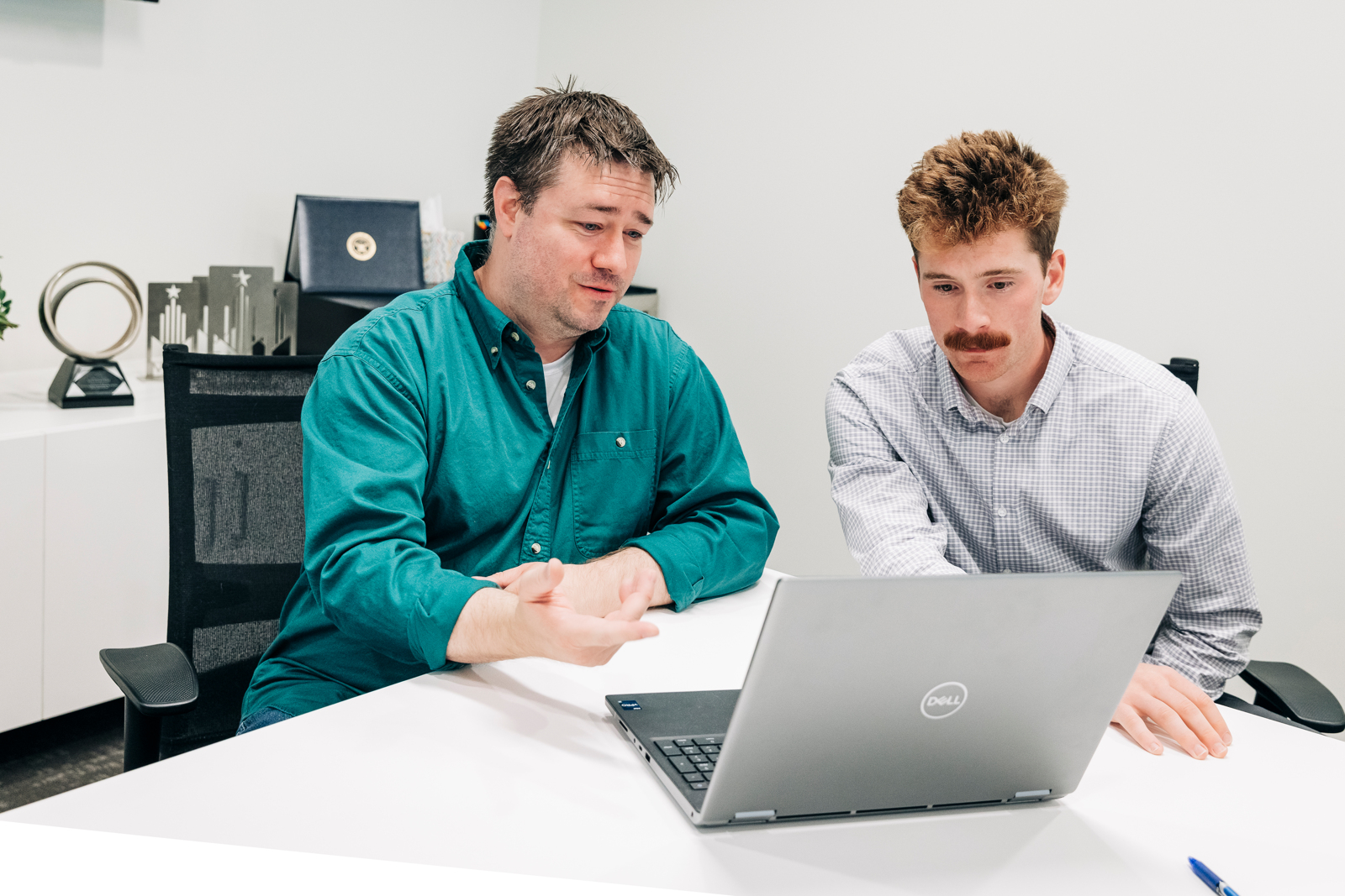 Two men sit at a white desk looking at a Dell laptop, discussing civil engineering plans. One gestures while the other points at the screen. Office items and awards are visible on a shelf in the background.