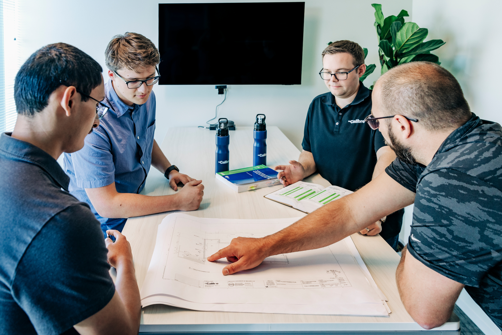 Four men, including a structural engineer, sit around a table with architectural plans, discussing and pointing at the drawings. Two blue water bottles and a tablet are on the table, with a TV and plant in the background.