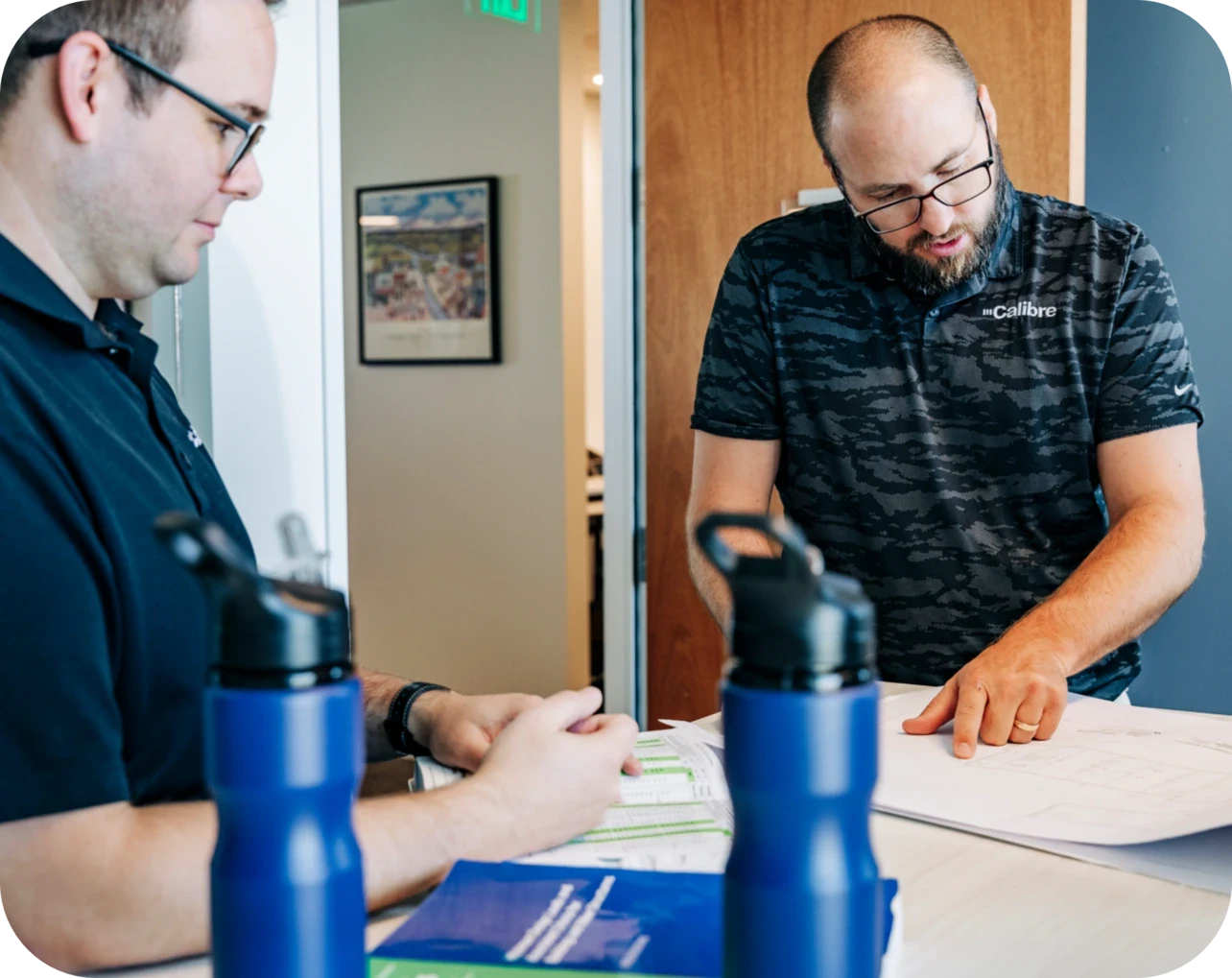 Two men discuss papers at a desk in an office, possibly reviewing colorado engineering plans. Both wear glasses and dark shirts. Two blue water bottles and blue folders are on the table as one points to documents while the other listens attentively.