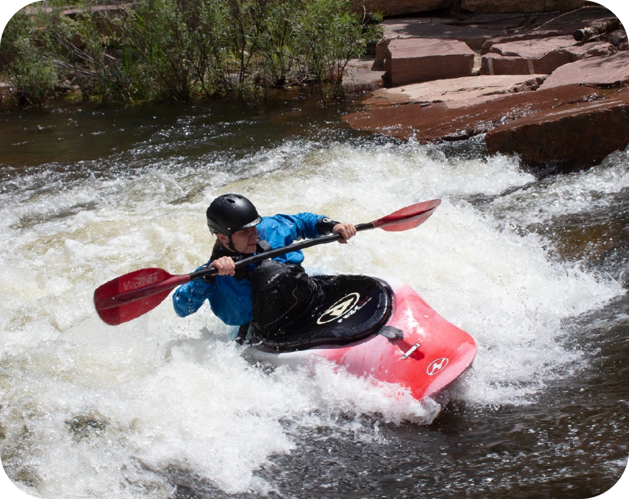 A person wearing a helmet and blue jacket, possibly a whitewater engineer, is kayaking through rapids in a red kayak, surrounded by rocky banks and greenery.