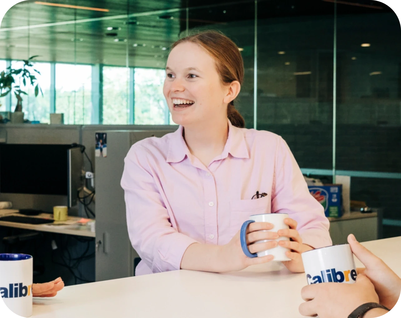 A person in a light pink shirt smiles while holding a white mug at a table in a modern office setting, interacting with others—perhaps discussing civil engineering projects or Colorado engineering opportunities.