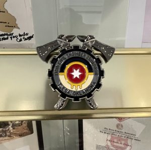 A military badge with a red star and yellow circle is displayed on a gold shelf in a glass case, surrounded by photographs, documents, and a patch labeled “IRAQ.”.