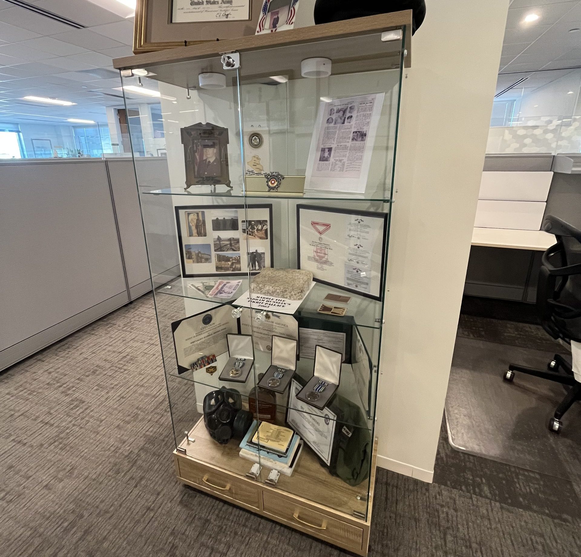 A glass display case in an office holds military memorabilia, including medals, certificates, photographs, a cap, a gas mask, and framed awards, all arranged on several glass shelves. Cubicles and desks are visible in the background.