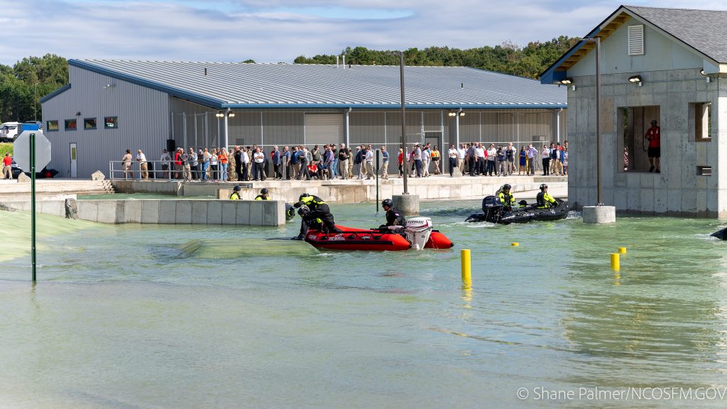 Several rescue teams in inflatable boats practice a water rescue exercise in a large outdoor training pool, while a crowd of people watches from behind a barrier near a gray building.