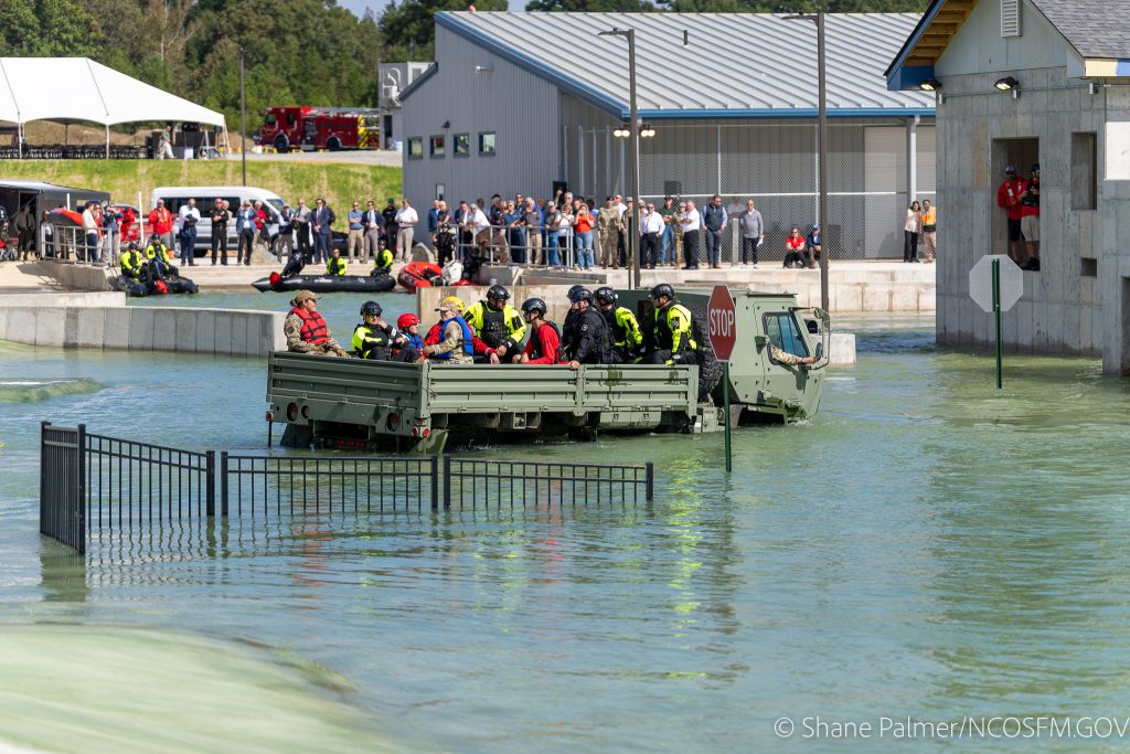 A military truck carrying rescue workers drives through floodwater at a training facility, passing a stop sign and partially submerged fence as onlookers observe from dry ground in the background.