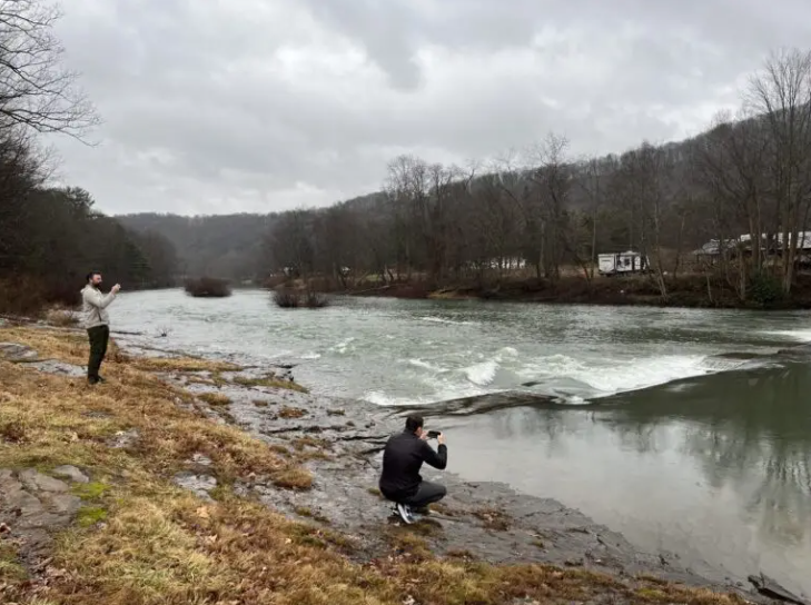 Two people are near a river on a cloudy day; one stands and gestures while the other crouches with a camera, capturing the scene. Bare trees and a few houses are visible in the background.
