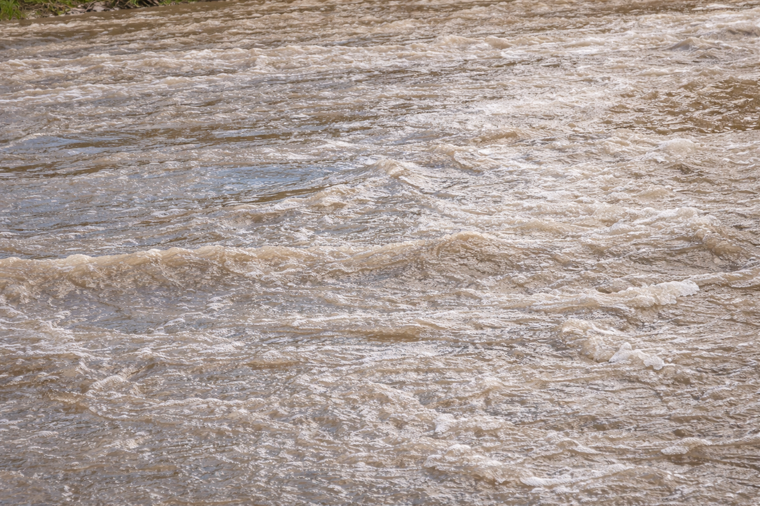 Fast-moving brown water with visible ripples and waves, indicating a turbulent river or stream. The surface appears foamy in some areas, suggesting strong currents and recent rainfall or flooding.