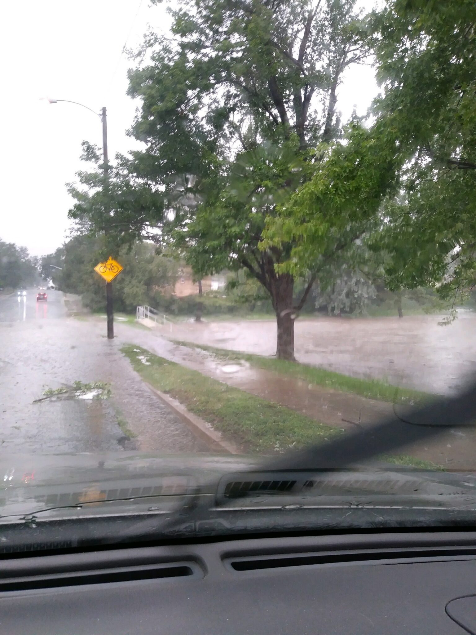 A view through a car windshield on a rainy day shows a flooded street. Water covers the road and sidewalk, with tree branches scattered and trees bending in the wind. A yellow bicycle crossing sign is visible.