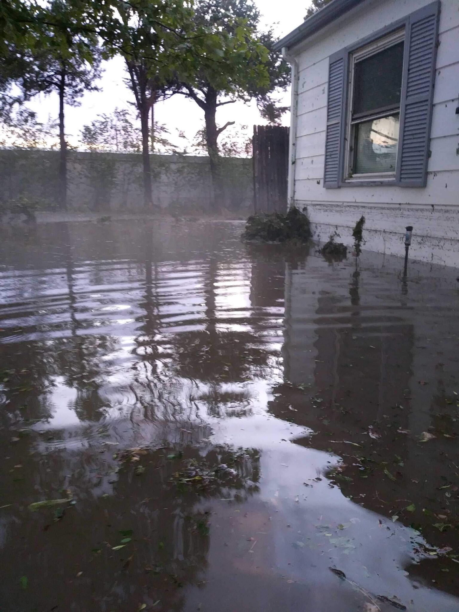 Floodwater surrounds the side of a house, nearly reaching the bottom of the window. Debris and muddy water reflect the house and nearby trees, indicating severe flooding in a residential area.