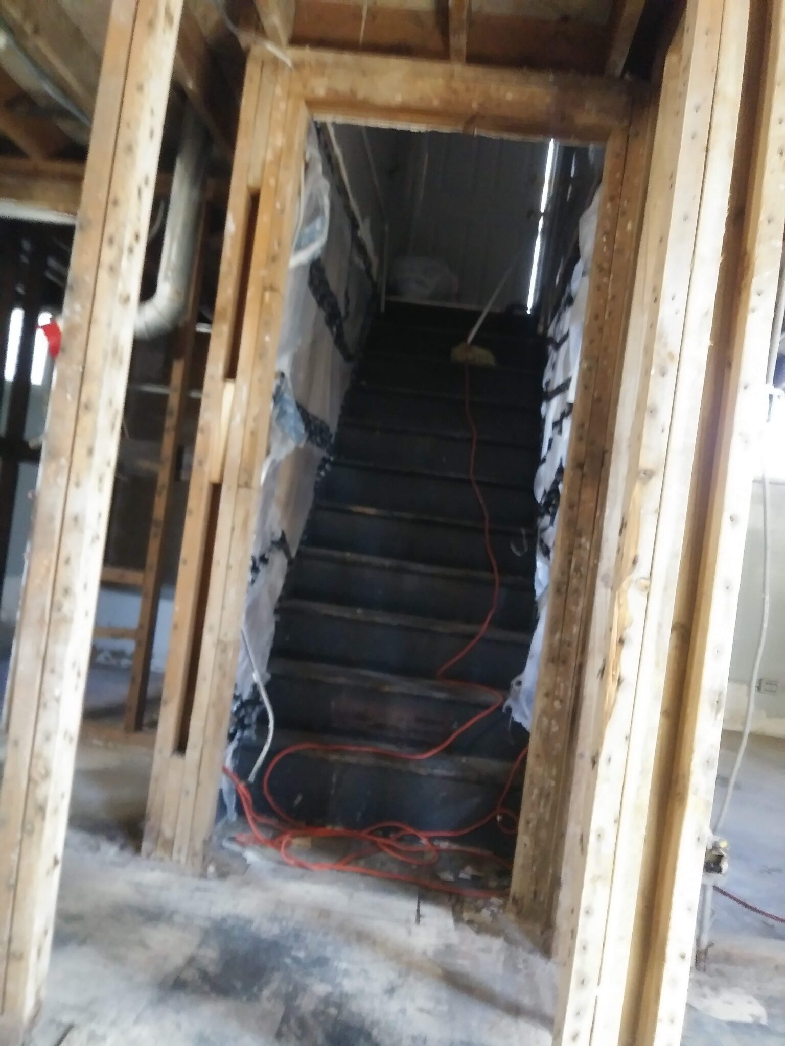 A set of dark wooden stairs in a house under construction or renovation, surrounded by exposed wooden beams and insulation, with an orange extension cord running up the steps.