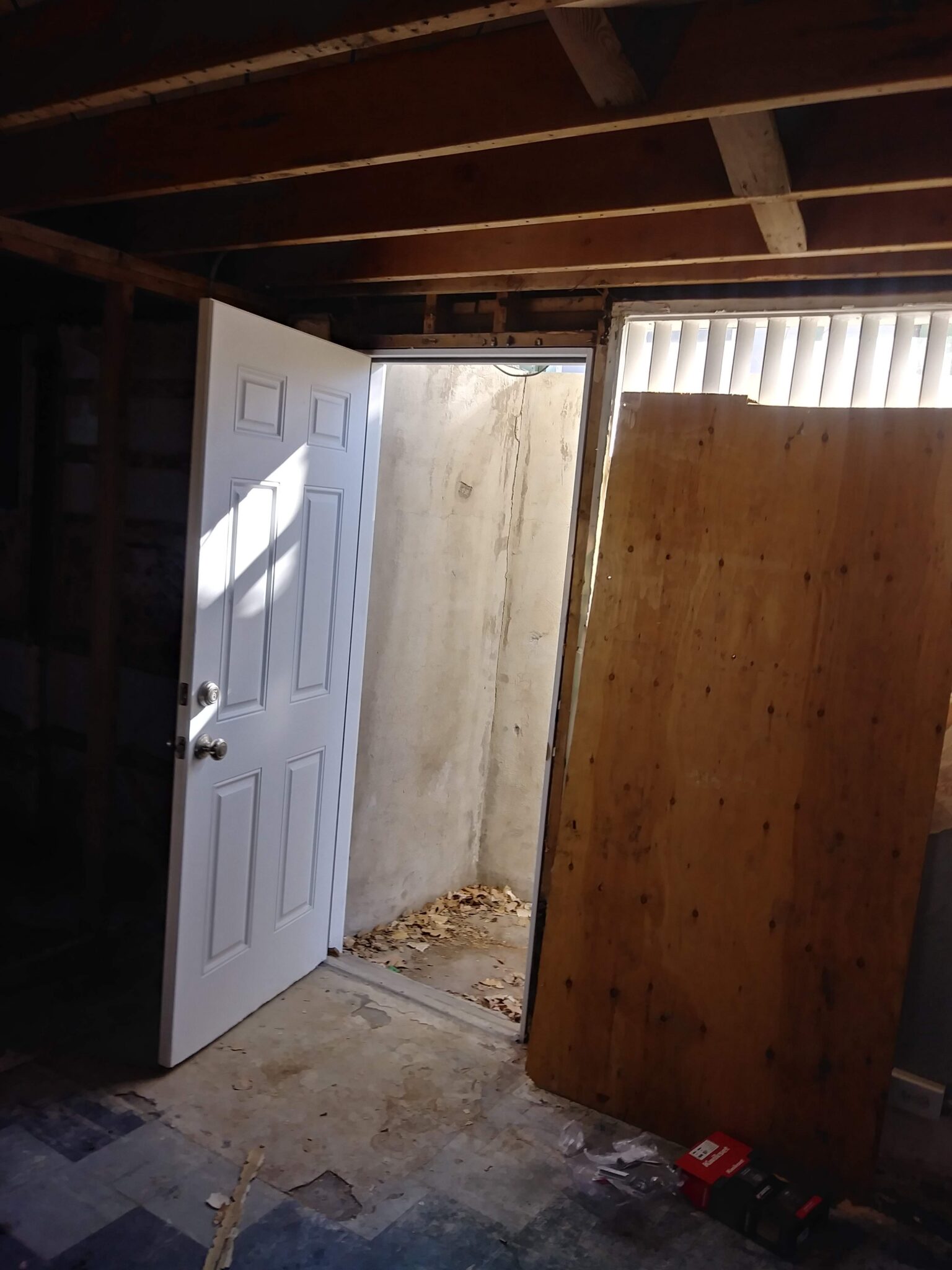 A partially open white door in a dimly lit room leads to a small concrete area with scattered leaves. A large wooden board leans against the wall beside the door, and unfinished ceiling beams are visible overhead.