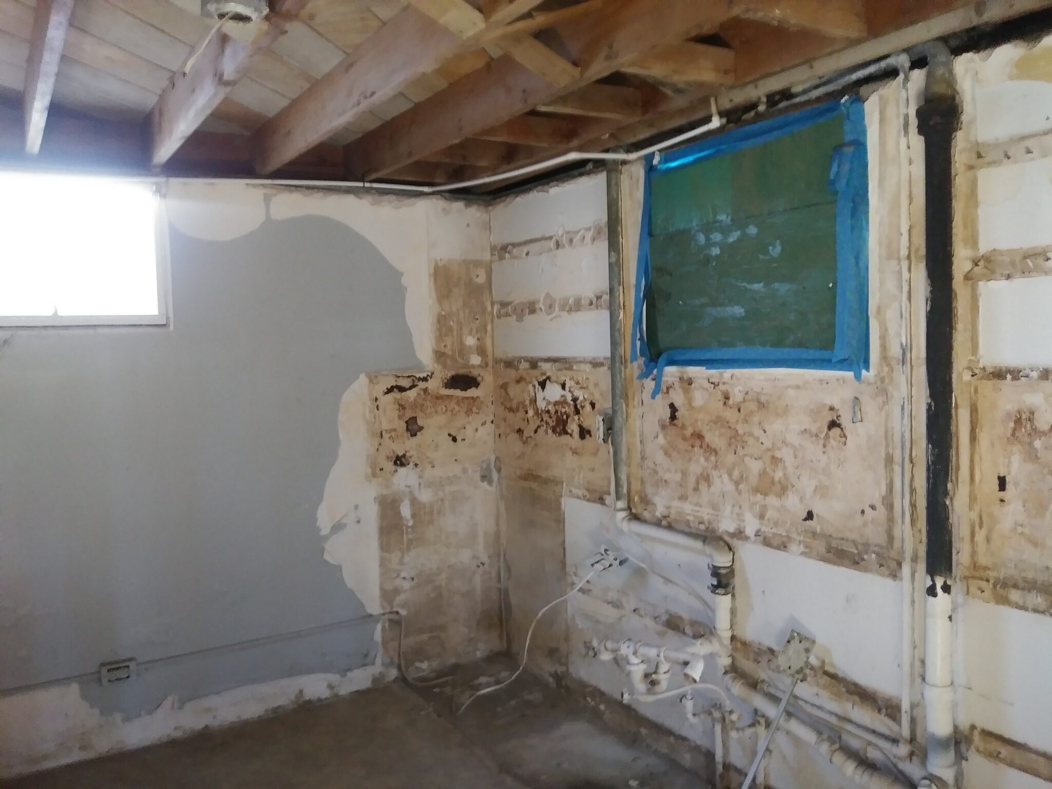 A partially demolished basement room with exposed wooden ceiling beams, peeling and damaged walls, visible plumbing pipes, and two windows—one taped over with blue plastic.