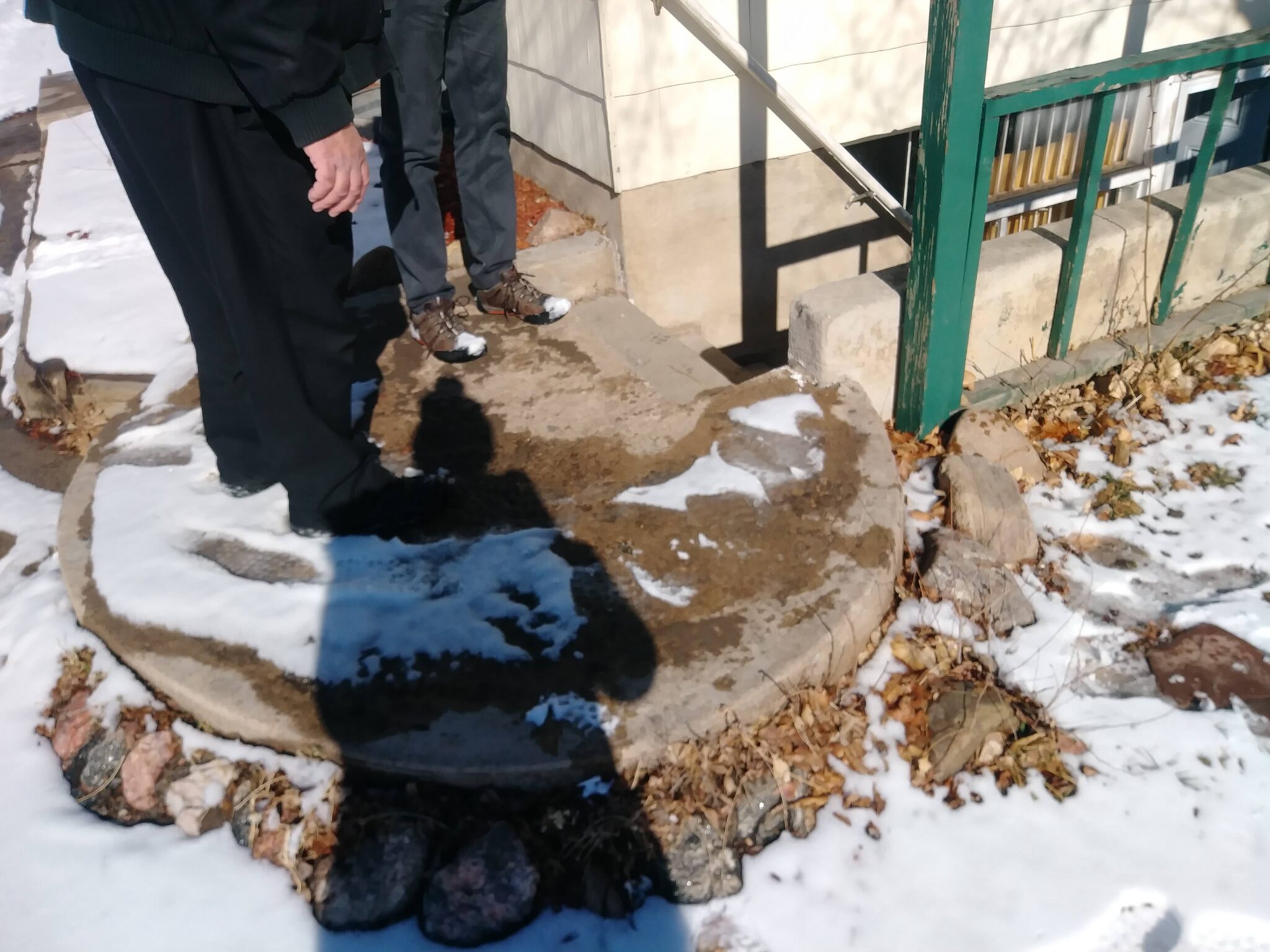 Two people stand outside on a snowy sidewalk near a round concrete area bordered by rocks. One persons shadow is visible on the ground. There is a green metal railing and building wall in the background.