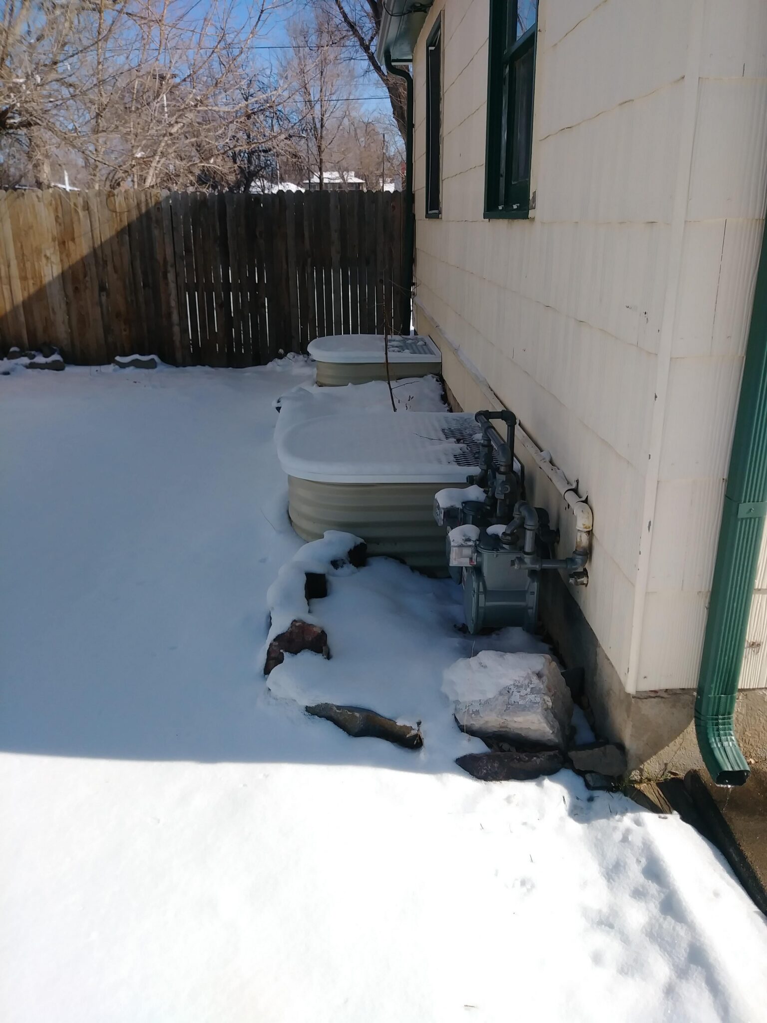A snowy backyard with a wooden fence, two raised garden beds, and a gas meter along the side of a house. Some rocks are partially buried in snow near the house. Leafless trees are visible in the background.