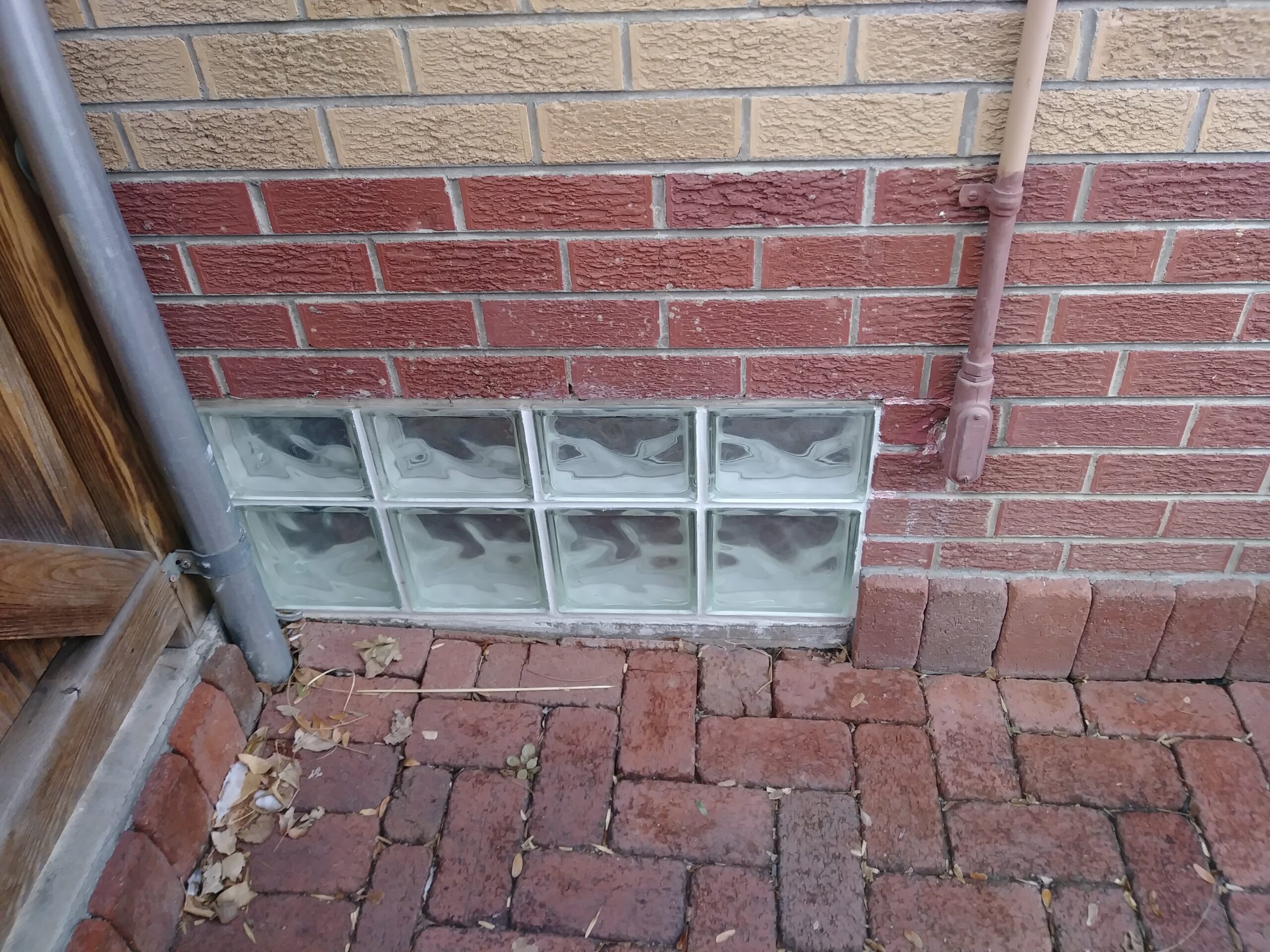 A small, rectangular glass block window set in a red brick wall near the ground. The window is bordered by a wooden fence on the left and a pipe running vertically along the wall. Brick pavers cover the ground.