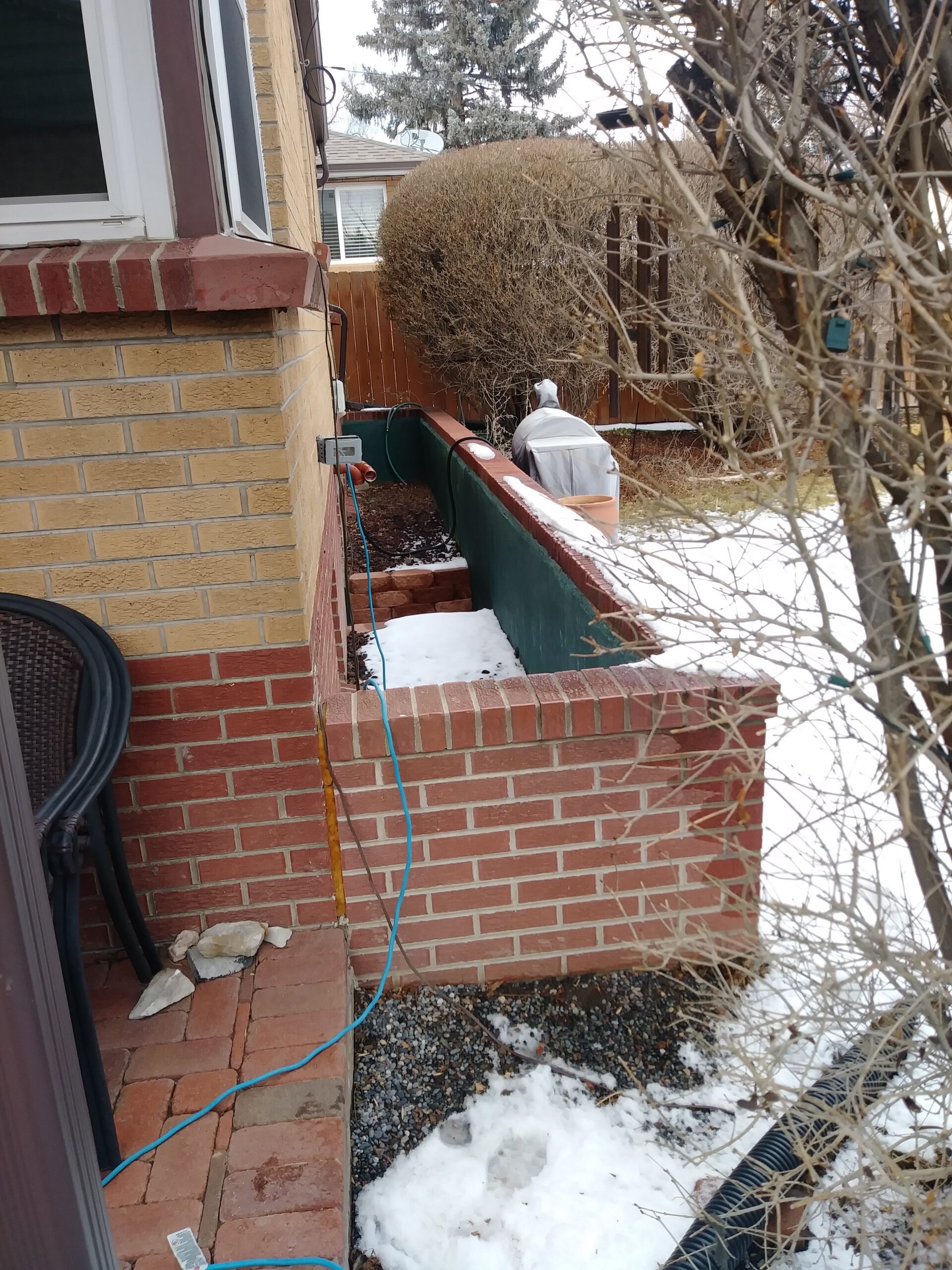A brick garden bed along the side of a house with some snow on the ground and in the bed, a green extension cord, patio chair, and leafless bushes are visible in a wintery backyard scene.