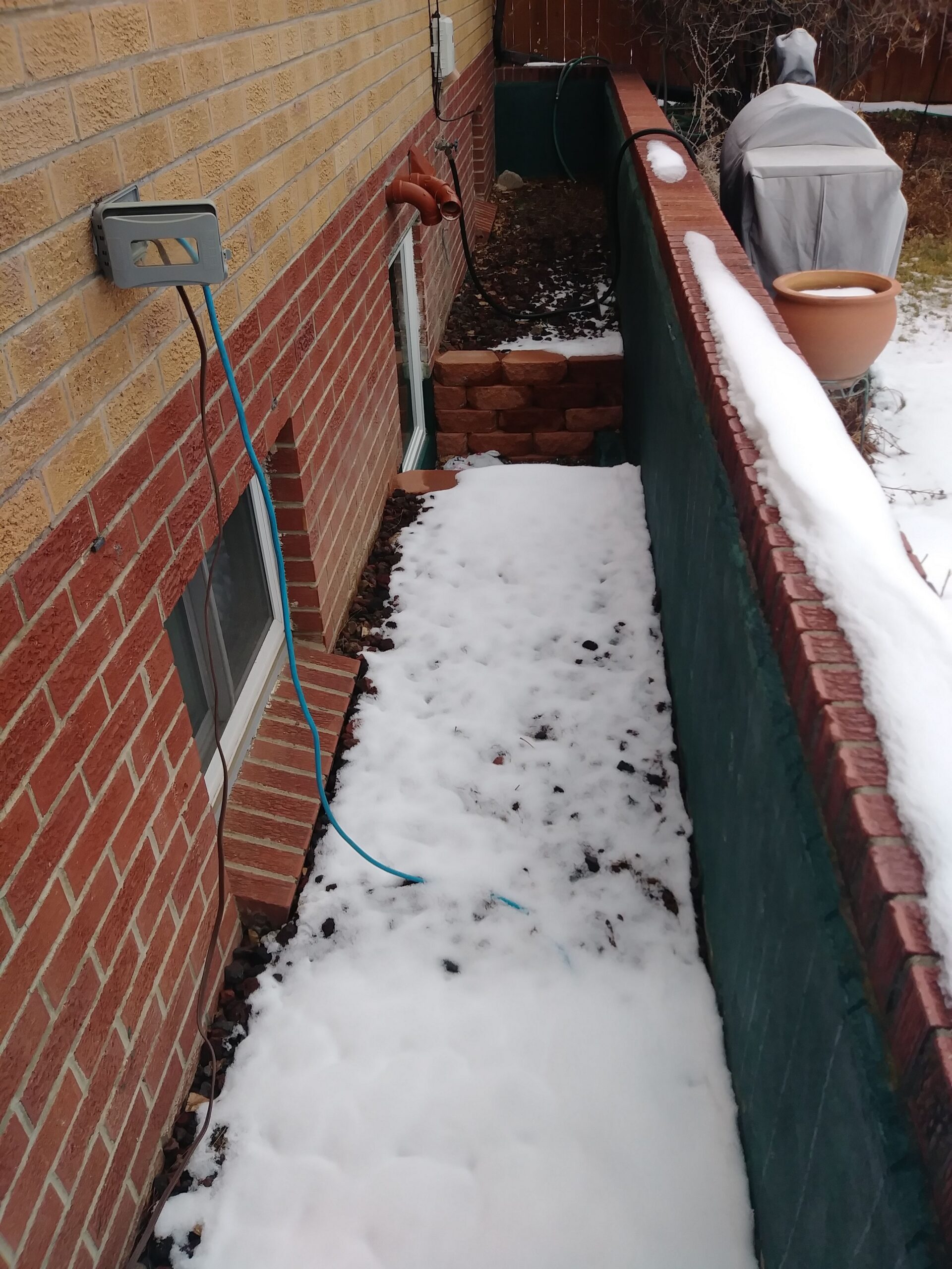 A narrow outdoor walkway next to a brick house is covered in patchy snow. A blue hose hangs from the wall, and a grill with a cover is in the background. A small retaining wall leads to a lower window.