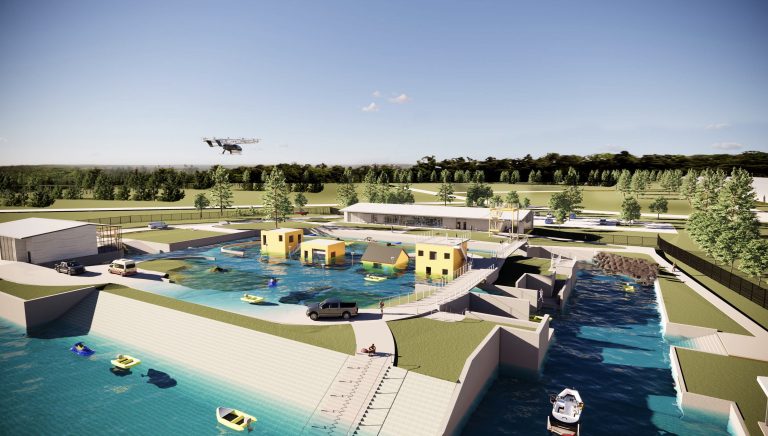 A modern water rescue training facility features pools, yellow buildings, vehicles, and boats, surrounded by greenery. A drone flies overhead under a blue sky with scattered clouds.