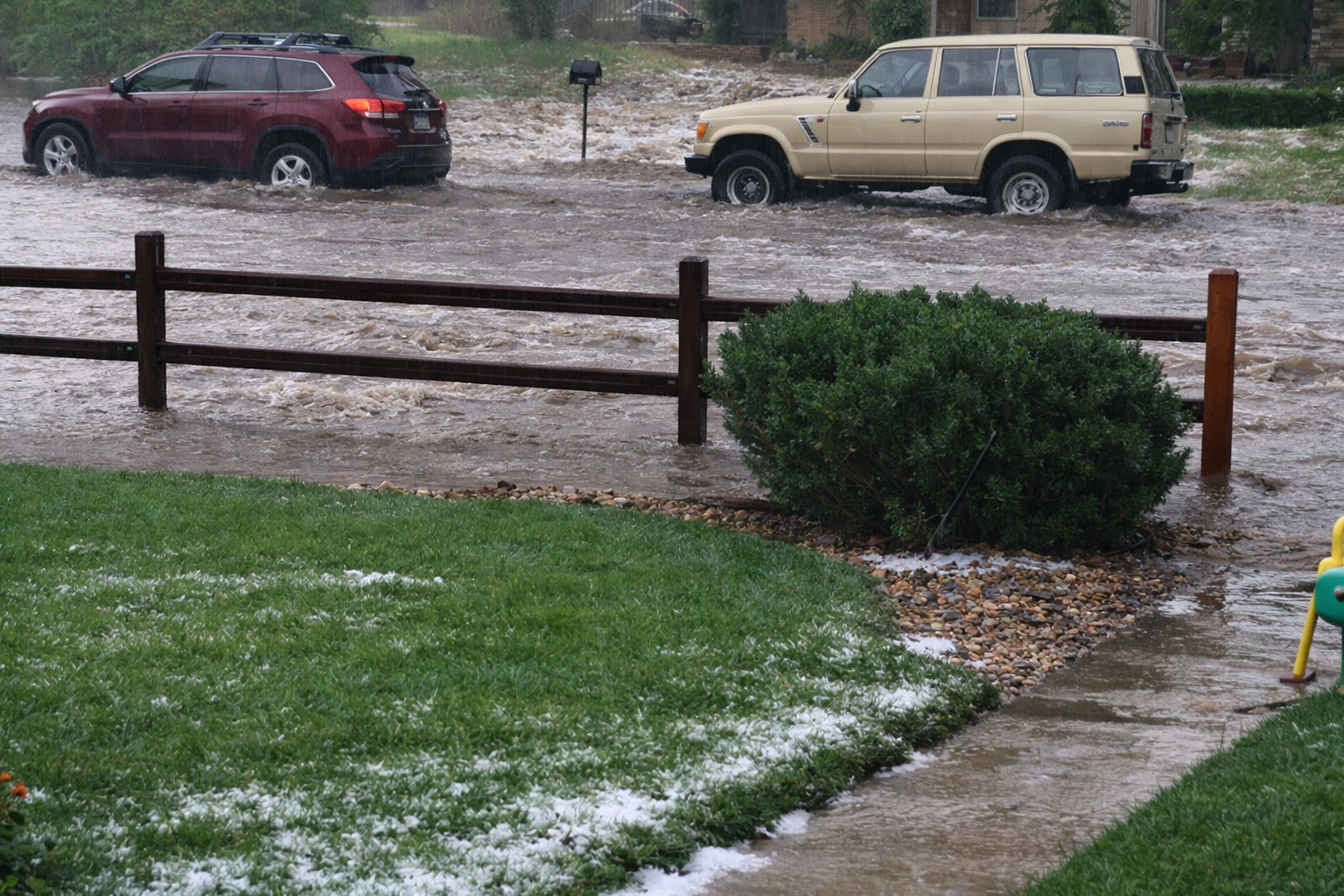 Two vehicles drive through a flooded street during heavy rain. A wooden fence and green lawn are in the foreground, with water flowing across the road and some hailstones scattered on the grass.