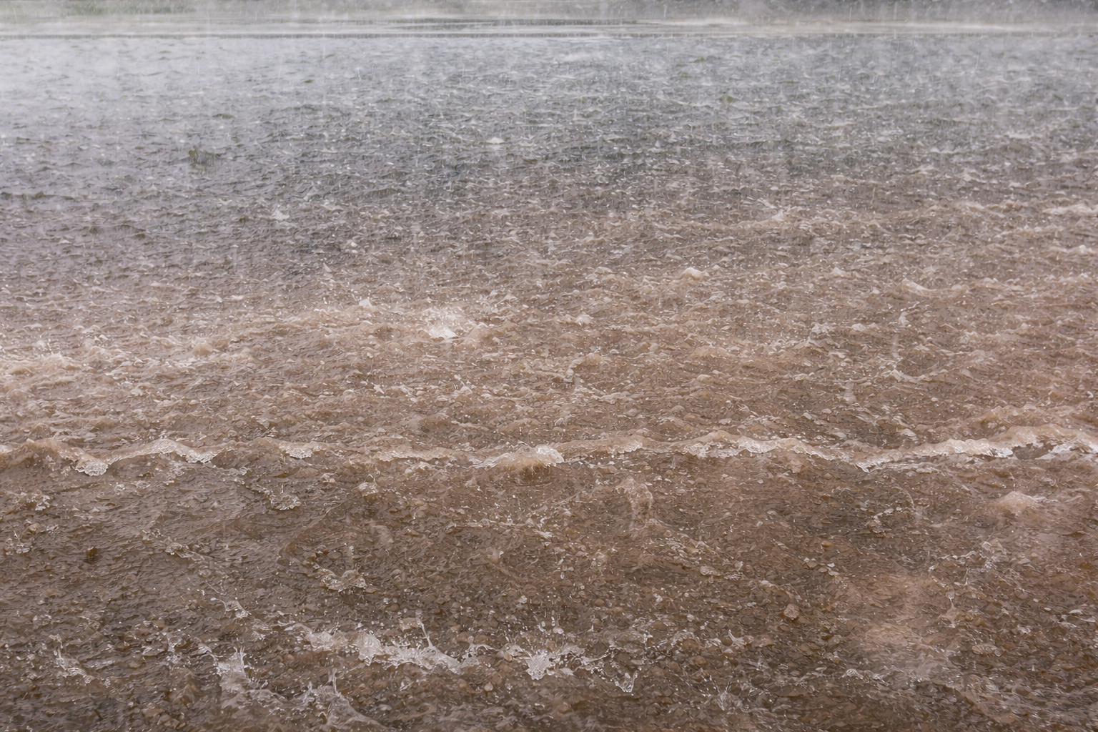 Heavy rain falling on a muddy surface, creating ripples and splashes across the waterlogged ground. The scene captures the texture of rain hitting the earth, with visible droplets and waves.