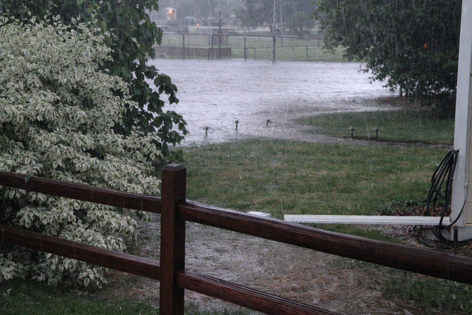 Rain pours down in a backyard, flooding the grass and parts of the patio. Wooden fencing, green bushes, and trees are visible, while the sky is gray and overcast, adding to the stormy atmosphere.