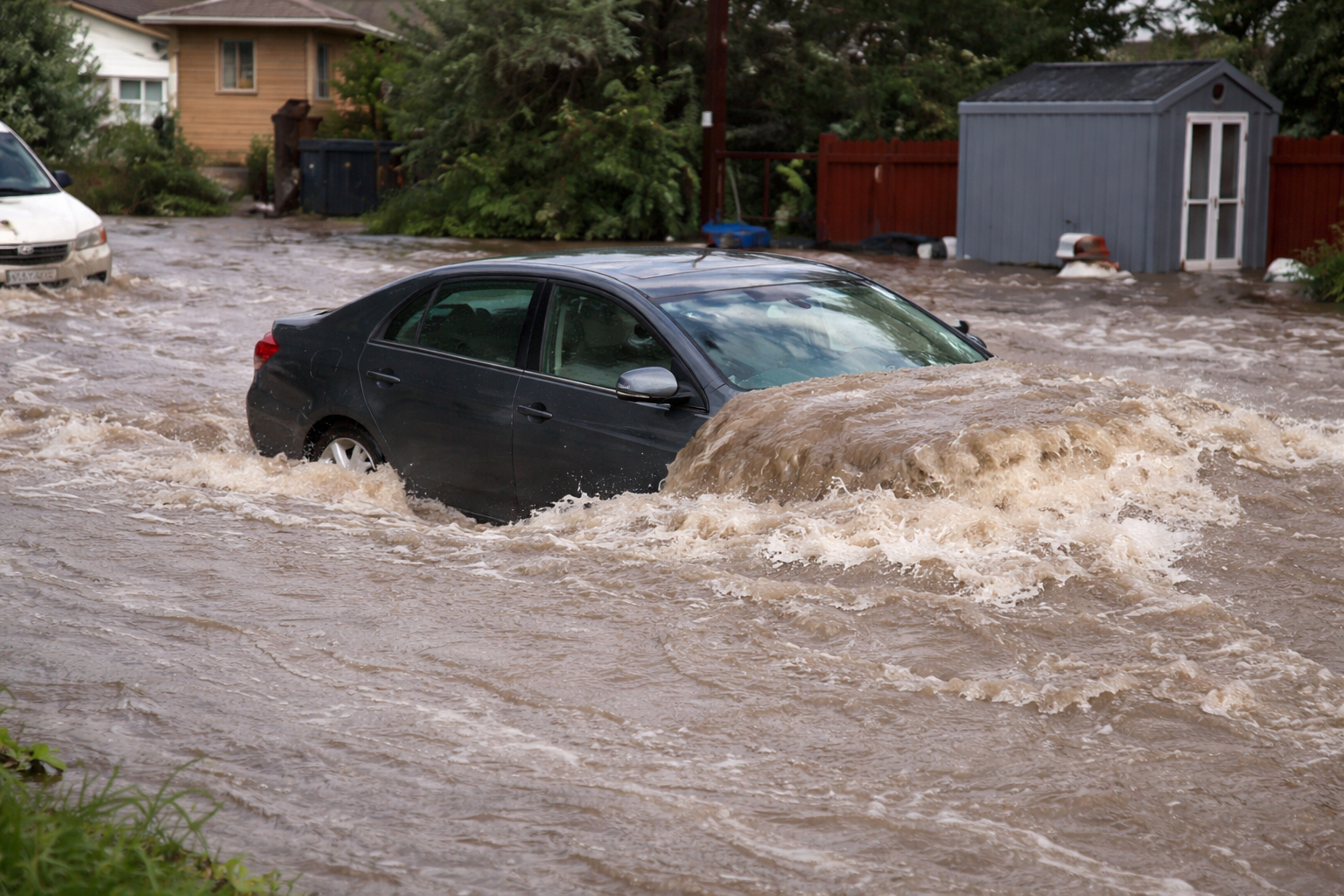 A black car is partially submerged and driving through a flooded residential street, with strong currents of muddy water surrounding it and nearby houses and sheds in the background.