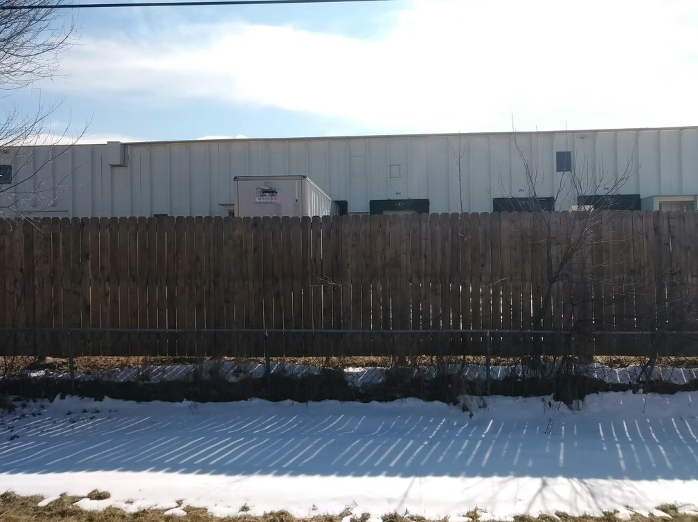 A tall wooden fence stands in front of a white industrial building. Snow covers the ground along the fence, with some bare patches visible. Shadows from the fence fall across the snow under a bright sky.