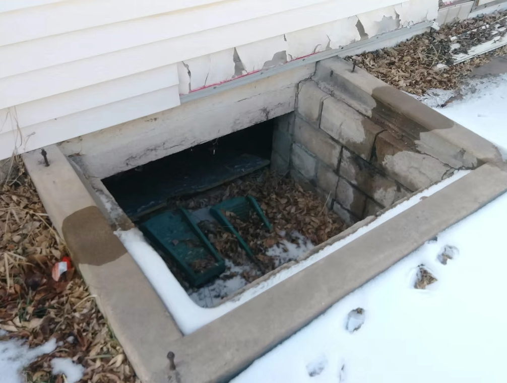 A basement window well against a house is partially filled with dried leaves and snow. Concrete borders the well, and the wall paint is chipped. Footprints and dead leaves are visible on the snowy ground nearby.