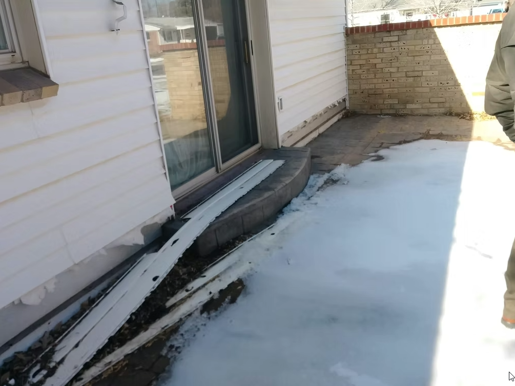 A patio area with dirty, melting snow covering the ground. Damaged siding panels are leaning against the wall near a sliding glass door, and a person is partially visible on the right side of the image.