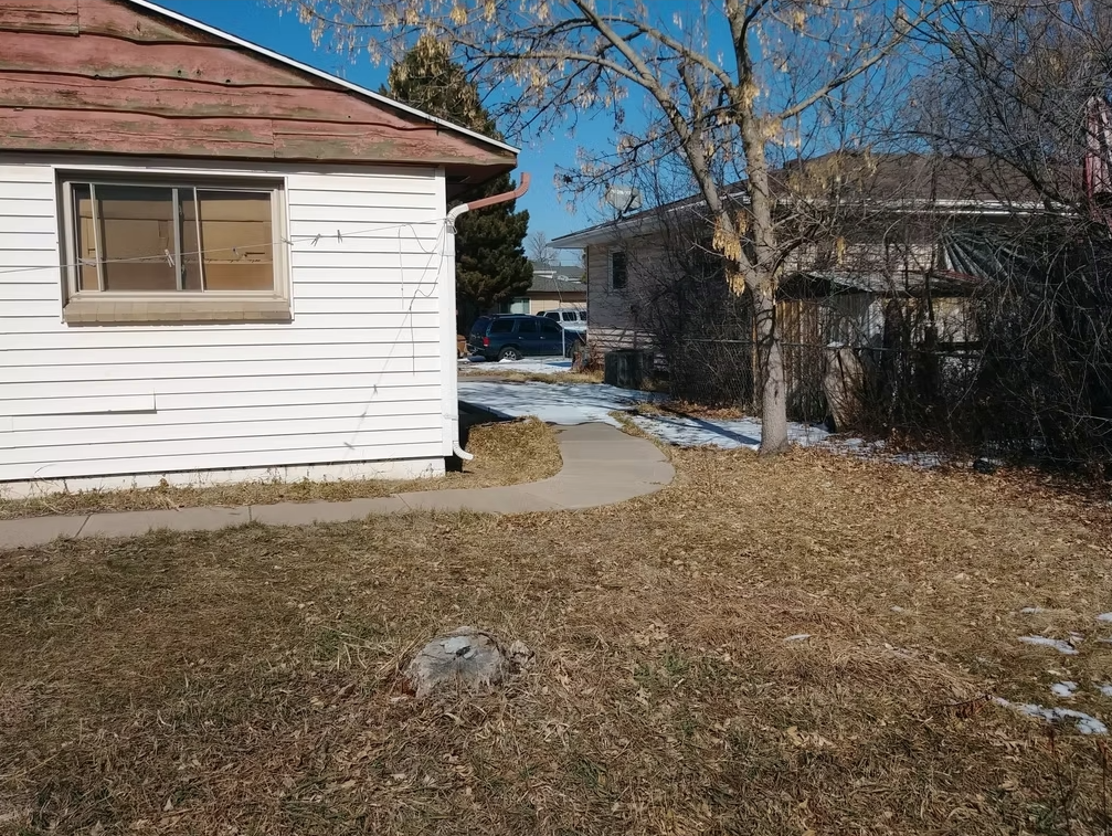 A yard with patchy brown grass, a bare tree, a small sidewalk, and scattered snow. Part of a white wooden house is on the left, and another house with cars parked between them is in the background.