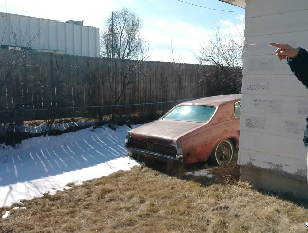 A rusty, old car is parked beside a house, with snow covering the ground near the car and dry grass in the foreground. A persons hand is visible, pointing from the right edge of the image.