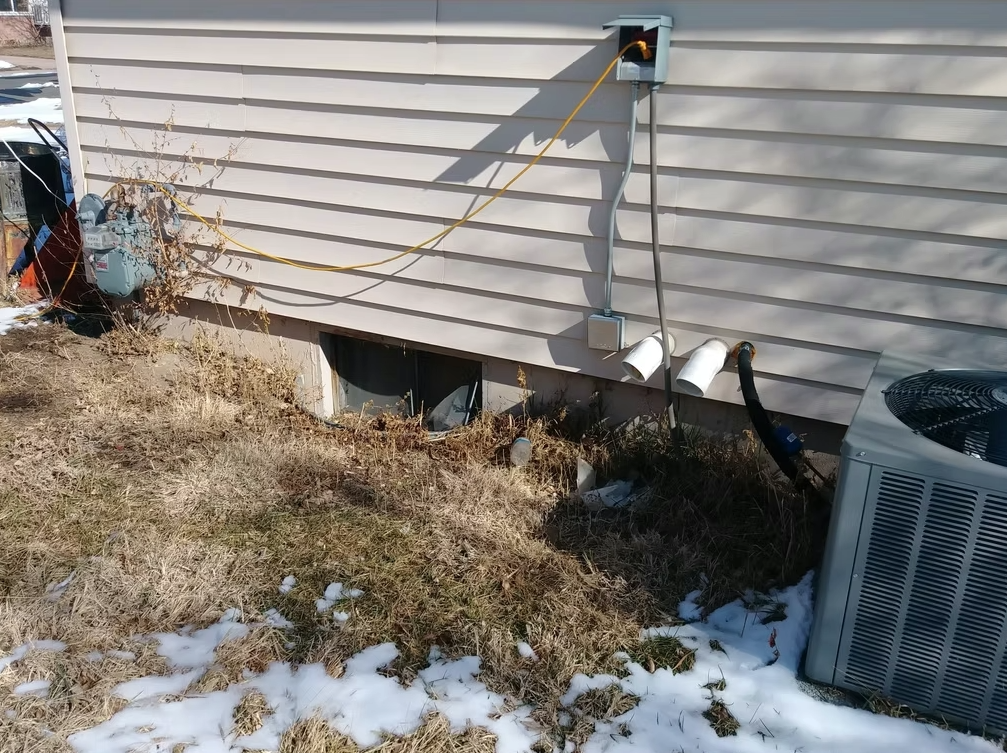 An outdoor HVAC unit sits beside a beige house. Electrical cords and pipes run into the siding. The ground is covered with dry grass, patches of melting snow, and some weeds near the house.