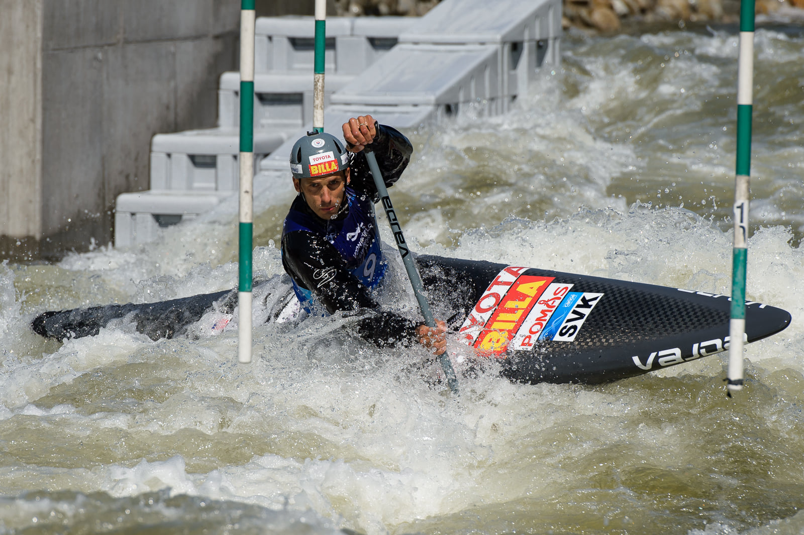 A male kayaker wearing a helmet and life vest maneuvers through turbulent whitewater, paddling past green and white slalom gates during a kayak competition.