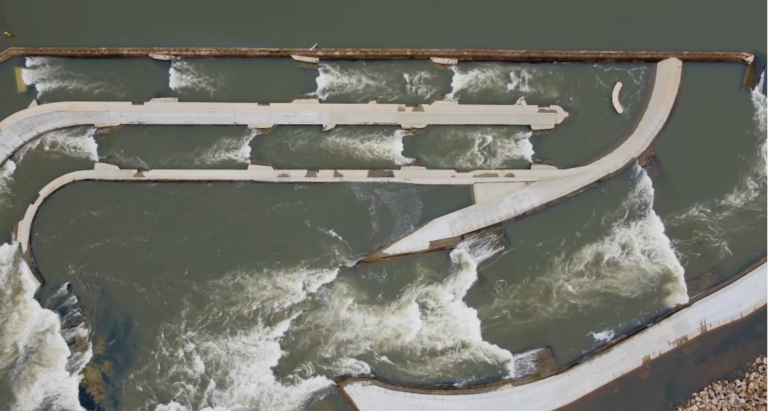 Aerial view of a concrete fish passageway or water channel structure, with water flowing rapidly through and around curved and straight barriers. White water turbulence is visible throughout the structure.
