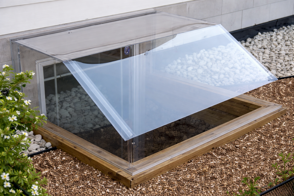 A clear plastic window well cover is partially open over a basement window, surrounded by white stones, gravel, and green plants near the house foundation.