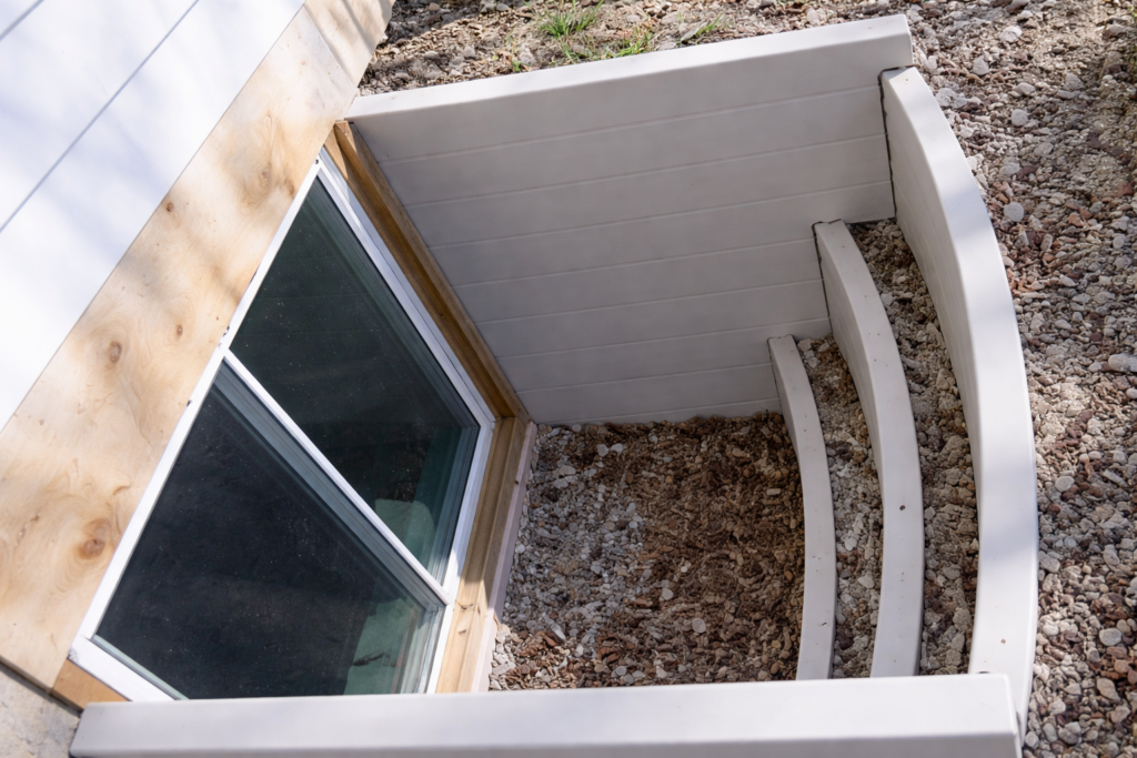 A basement window well with white retaining walls, gravel, and some dry leaves at the bottom, next to a wooden house exterior.
