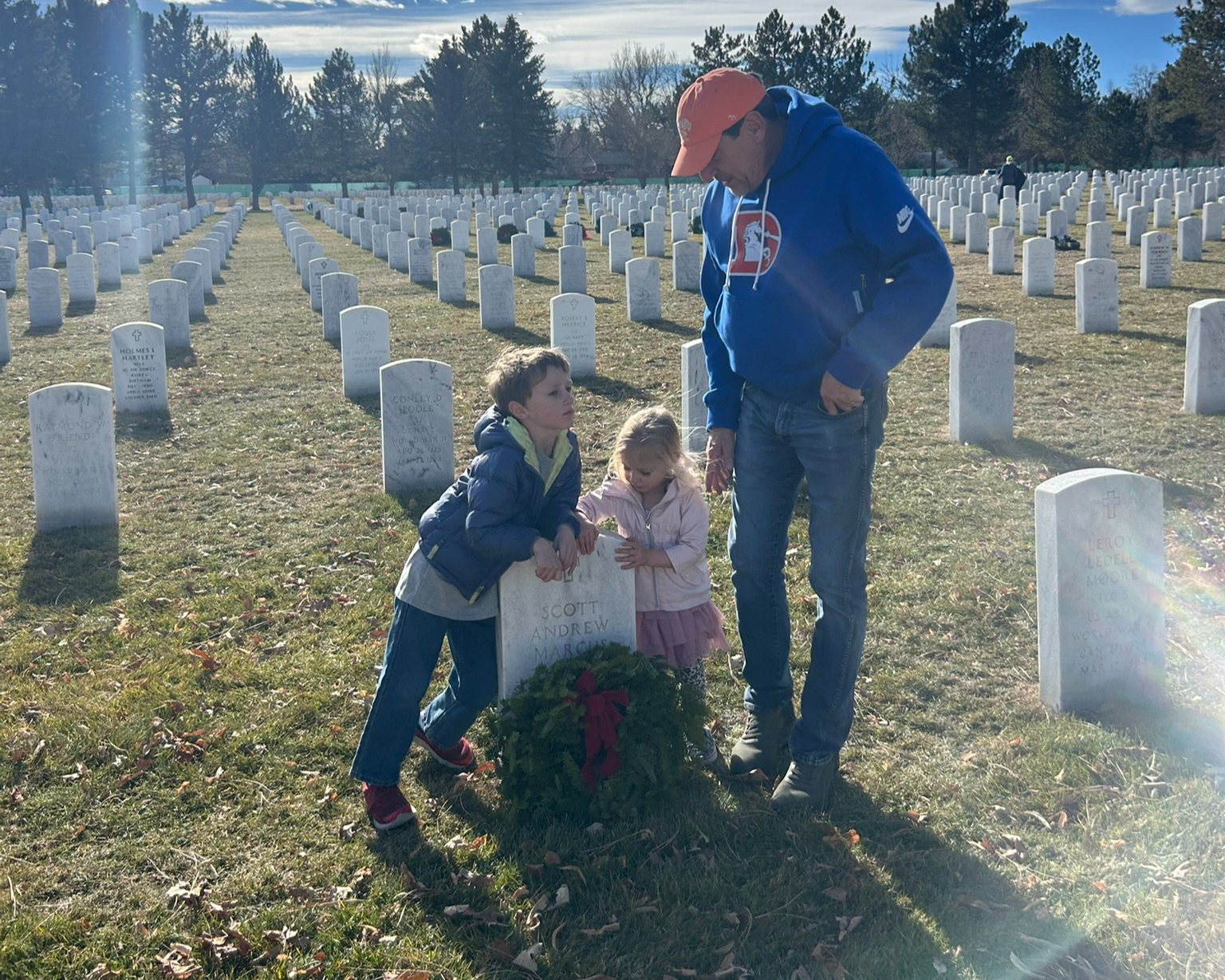 A man and two children stand by a gravestone with a wreath in a cemetery on a sunny day, surrounded by rows of white headstones and trees in the background.