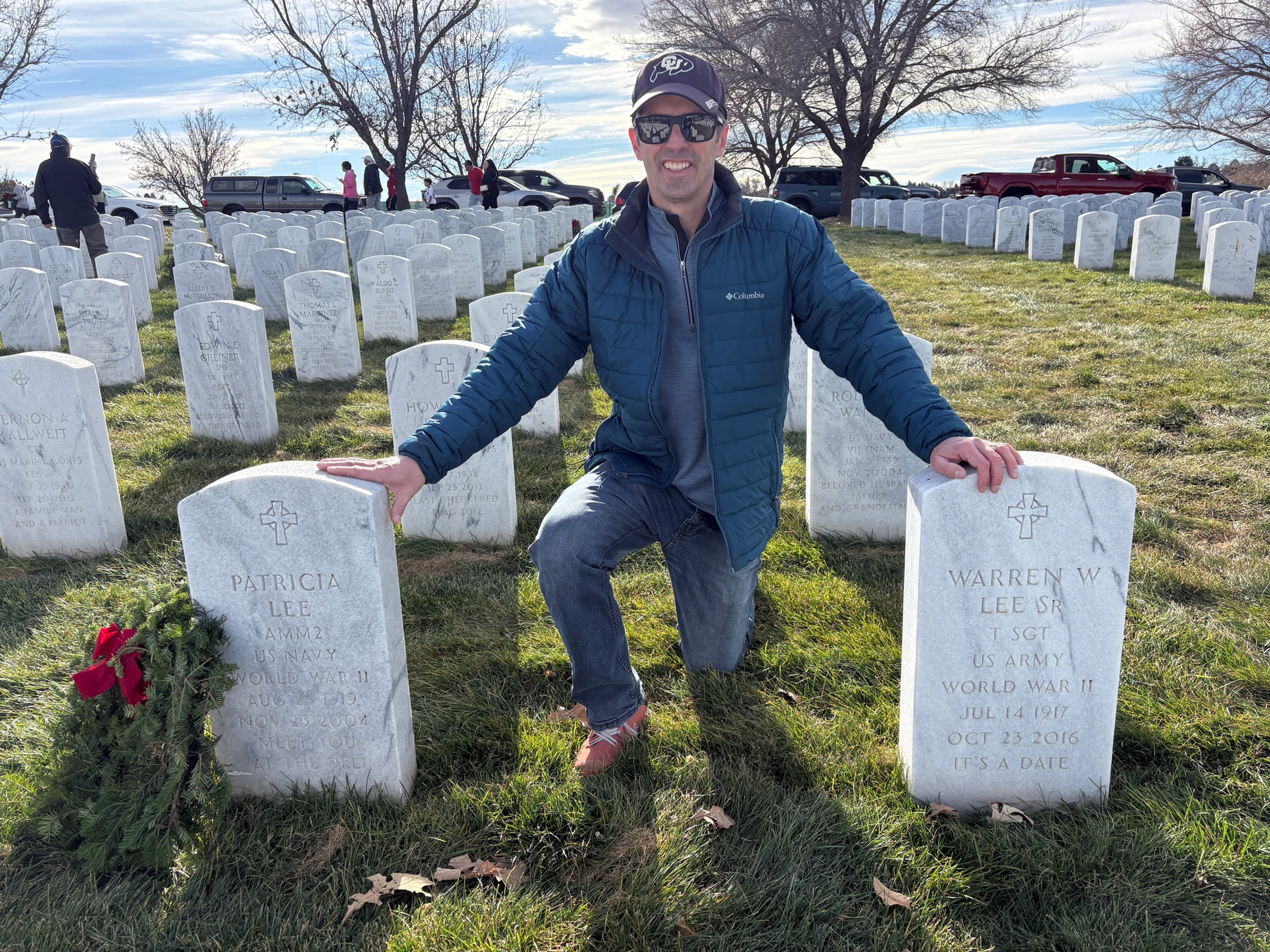 A man in sunglasses and a blue jacket kneels between two white gravestones at a cemetery on a sunny day, with rows of similar headstones and several cars in the background.