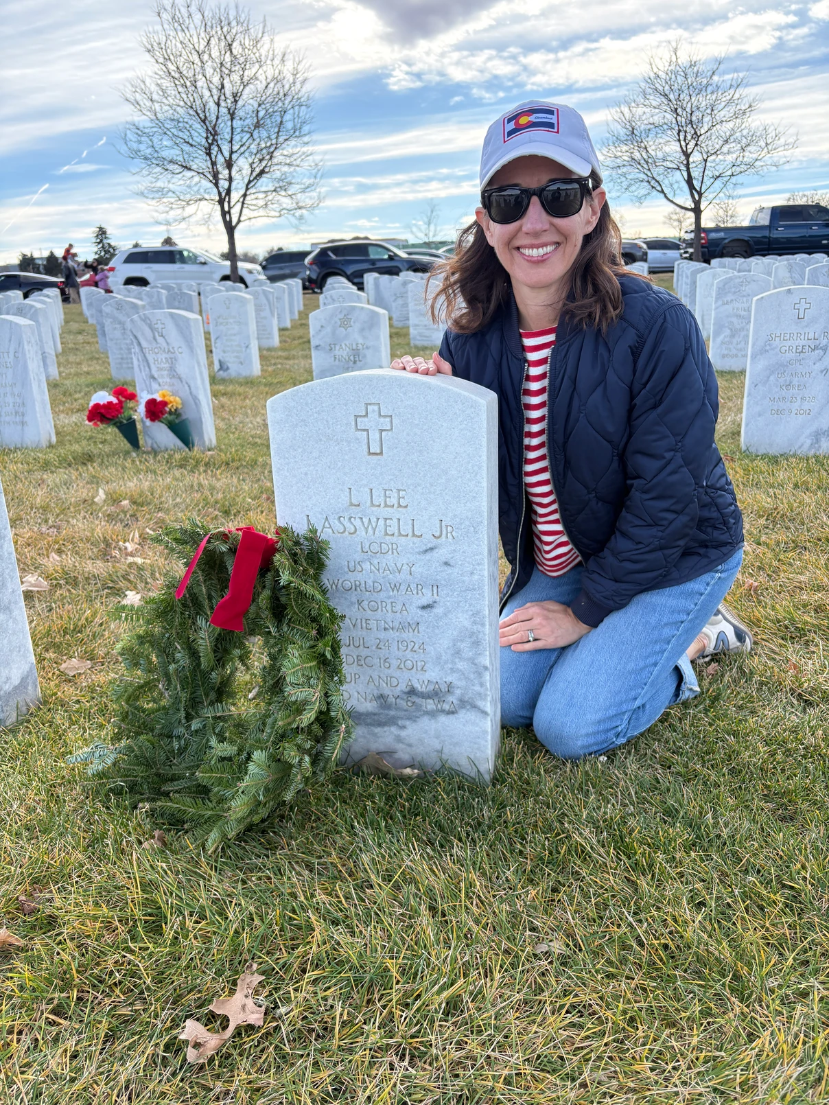 A woman wearing sunglasses, a striped shirt, navy jacket, and baseball cap kneels by a decorated military gravestone in a cemetery, smiling, with a wreath placed on the headstone. Other graves and flowers are visible.