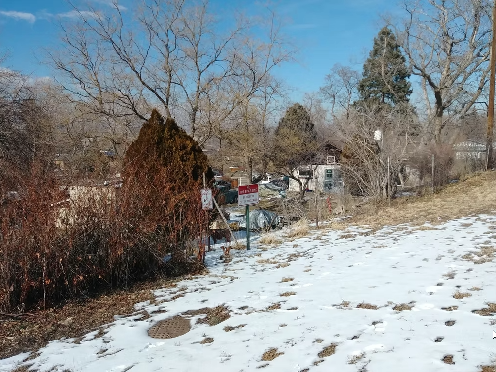 A partially snow-covered yard with leafless trees, brown bushes, and scattered buildings and vehicles in the background under a clear blue sky. A red sign and a manhole cover are visible in the foreground.