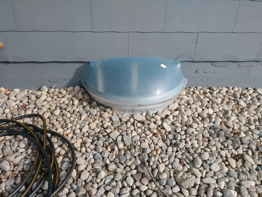 A translucent blue window well cover sits on a bed of light-colored rocks next to a gray building wall. A coiled black hose lies on the rocks in the lower left corner.