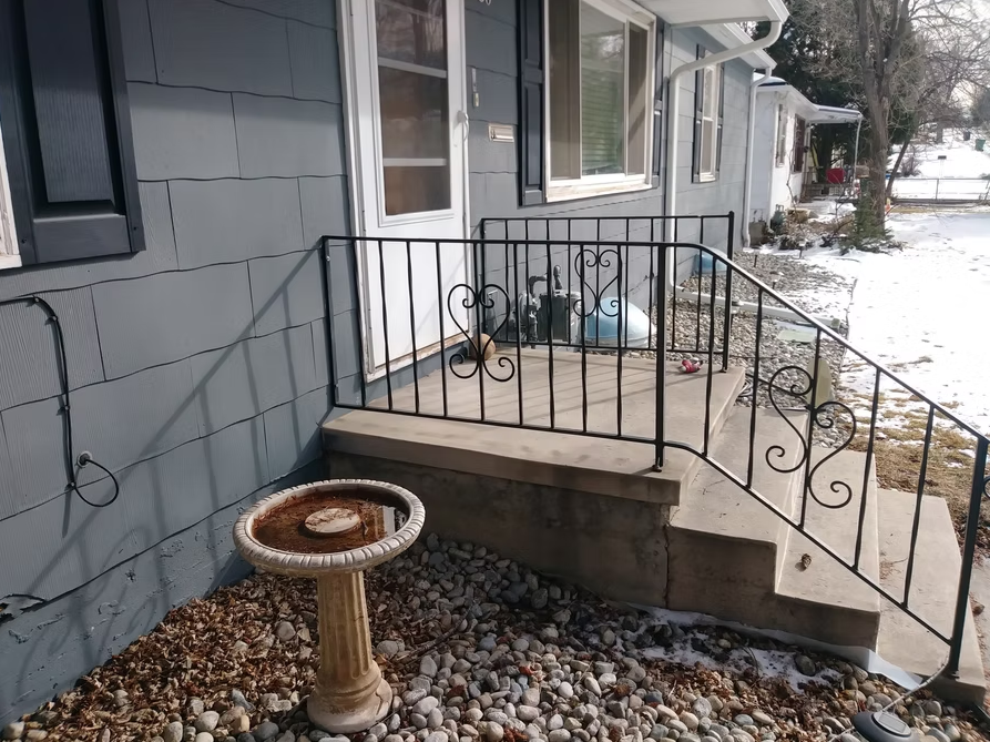 A small concrete porch with black metal railings leads to a white door on a gray house. A weathered birdbath stands in front, surrounded by rocks and dry leaves; some snow is visible on the ground and roof.
